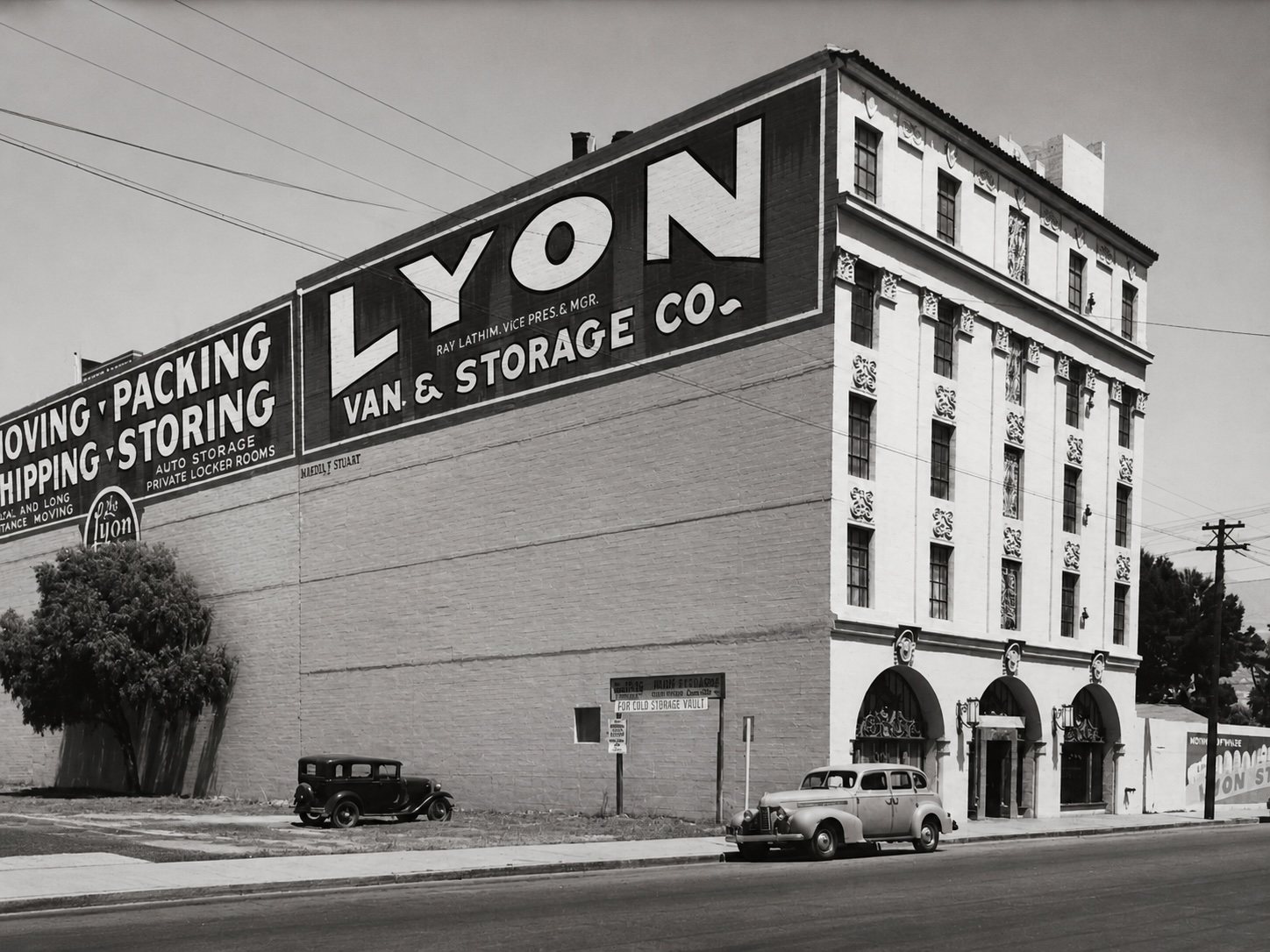 Historic photograph of the Lyon Van & Storage Co. building from Cota Street, its upper flank painted with a large sign reading Moving, Packing, Shipping, Storing, with period automobiles at the kerb.