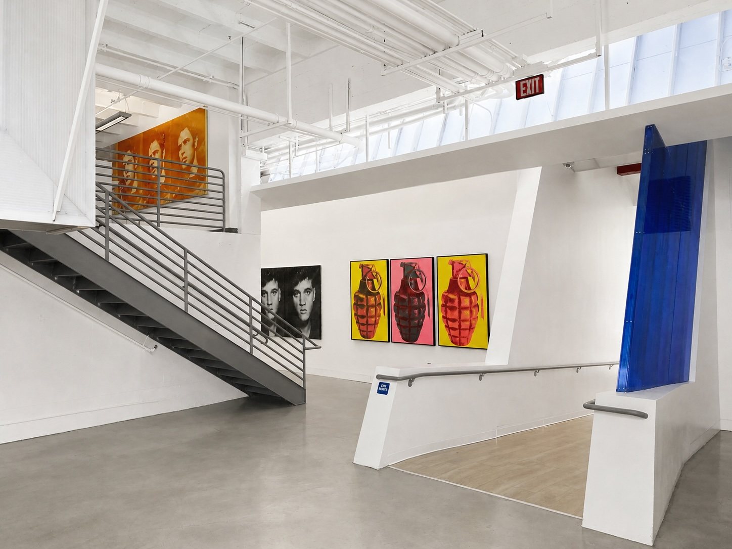 Suite 100: a double-height gallery volume with an open steel stair, clerestory daylight and large-format artwork on white walls.