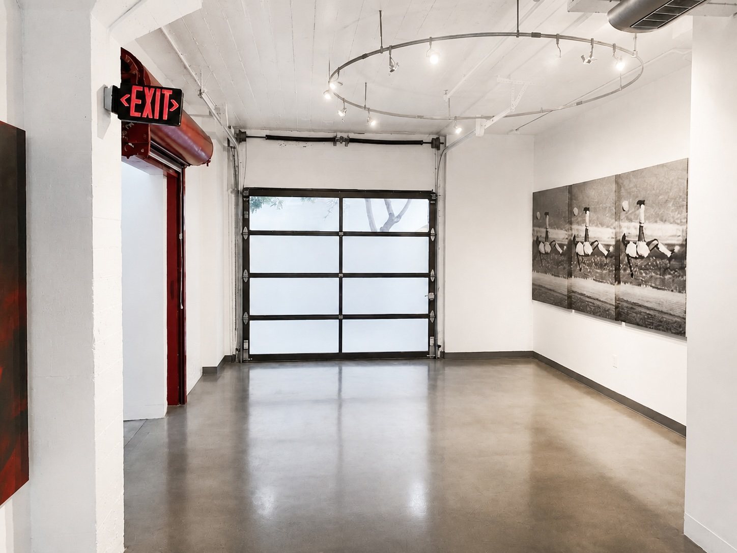 Suite 100 gallery room with a glazed roll-up door, circular track lighting and a vintage exit sign.